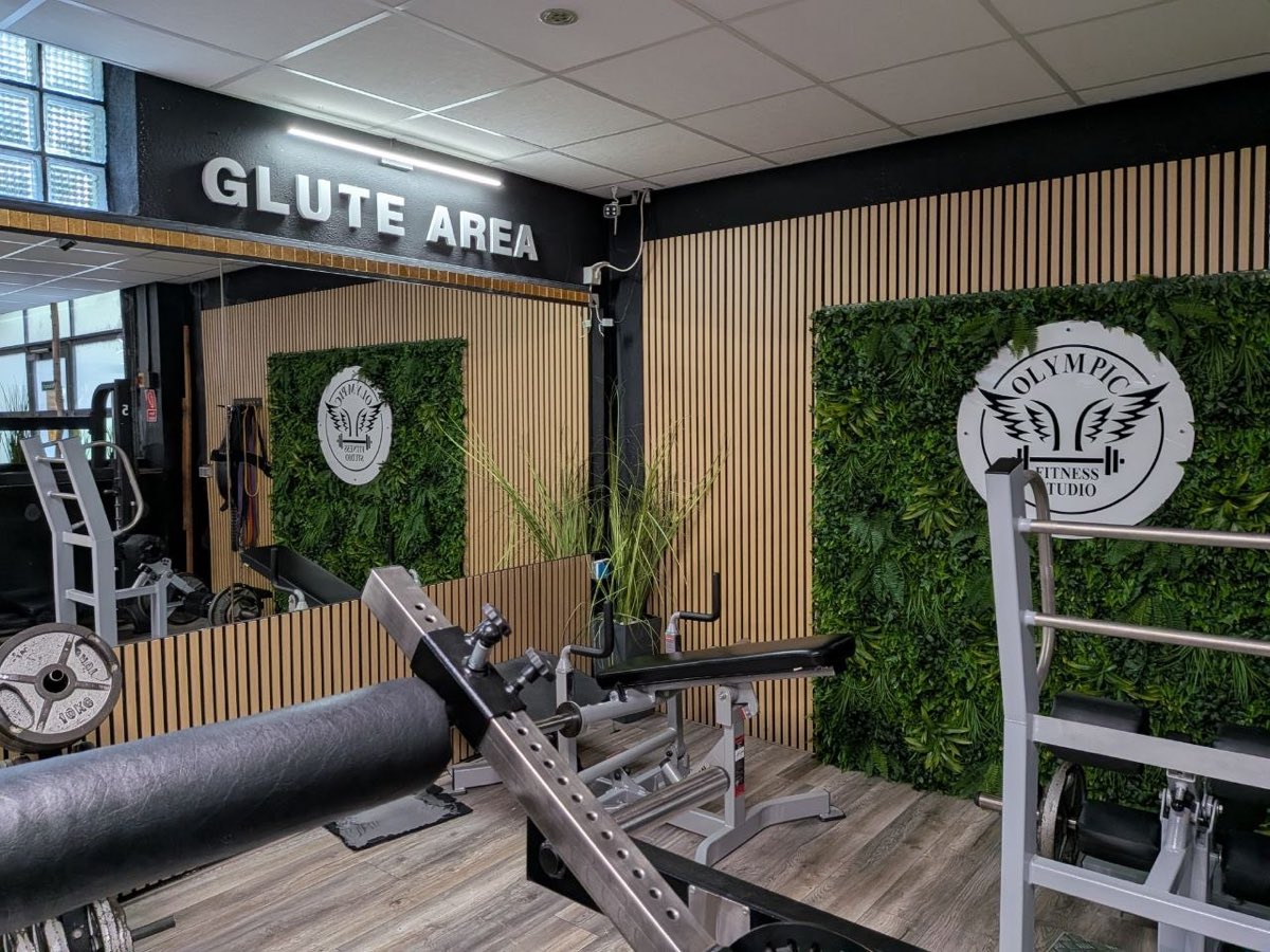 Glute Area mit Geräten vor begrünter Wand und Olympic-Gym-Logo